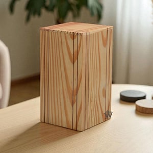 May include: A rectangular wooden box in light tones, featuring vertical grooves and a small logo. The box sits on a light-coloured table. A vase of dried flowers and coasters are in the background.