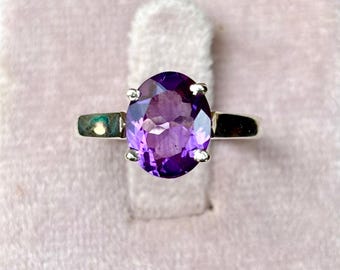 May include: A silver ring featuring a large, oval-cut amethyst gemstone. The deep purple stone is set in a classic four-prong setting, showcasing its vibrant color. The ring is displayed on a soft, pink velvet surface, highlighting the jewelry's elegance.