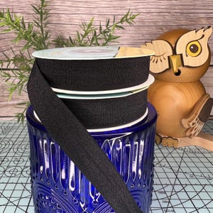 May include: Three spools of black elastic fabric ribbon are stacked on top of each other. The spools are sitting on a blue glass jar with a decorative pattern. The spools are wrapped in clear plastic with a white label.