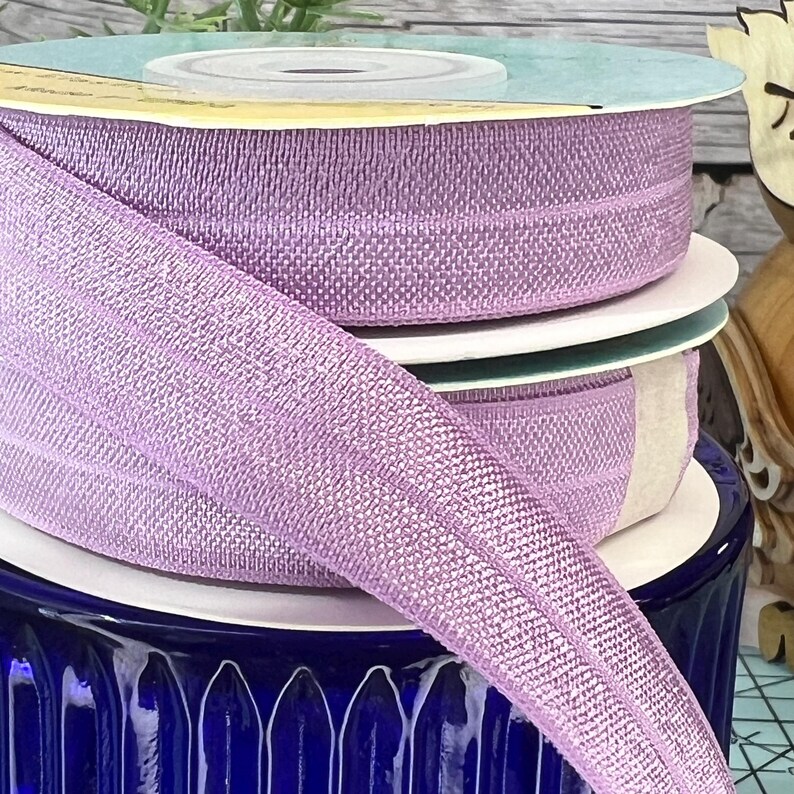 May include: A close-up of a roll of lilac-colored elastic ribbon with a shimmery finish. The ribbon is wrapped around a spool and is sitting on a blue and white ceramic base.