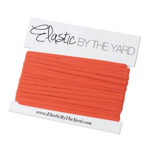 May include: A spool of orange elastic cord, wound on a white card with the text "Elastic By The Yard" and the website address "www.ElasticByTheYard.com".