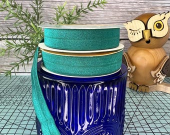 May include: Three spools of teal green elastic ribbon stacked on top of each other. The ribbon is sitting on a blue glass jar with a decorative pattern. The jar is sitting on a blue and white checkered surface.