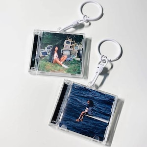 May include: Two square clear plastic keychain cases featuring miniature album cover art. One keychain displays a person sitting in a field with old computer monitors, while the other shows a person sitting on a diving board over deep blue ocean water.