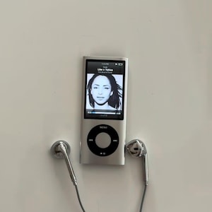 May include: A silver digital music player with a rectangular screen displaying a black and white portrait of Sade and the text Like a Tattoo. The device features a circular black click wheel with a central menu button and two silver wired earbuds.