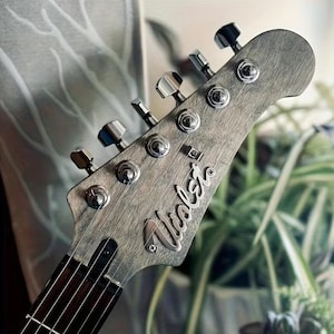 Könnte beinhalten: Ein Gitarrenkopf mit einer verwitterten grauen Holzmaserung und sechs chromfarbenen Stimmwirbeln an der Seite. Das Wort Violet ist in einer silbernen Schreibschrift auf der Vorderseite des Kopfes zu sehen, der in ein dunkles Griffbrett übergeht.