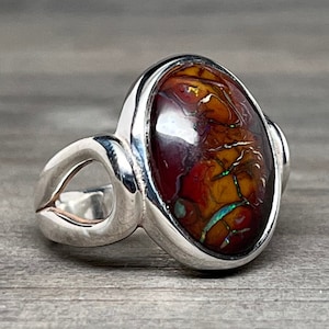 May include: Oval-shaped silver ring featuring a vibrant opal stone. The opal displays a mix of red, orange, and yellow hues with iridescent flashes of green and blue. The ring's band has a unique, open design.