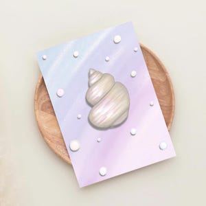 May include: A digital illustration of a pearlescent seashell on a pastel background of blue and purple hues. White, circular accents are scattered around the shell. The artwork is presented on a wooden plate.