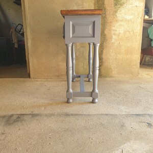 May include: A small, gray painted wooden side table with a dark brown top. The table has four turned legs and a rectangular cabinet door. The table is approximately 30 inches tall.