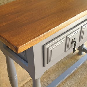 May include: A wooden console table with a stained brown top and a gray painted base. The table features three drawers with decorative molding and a keyhole. The legs are turned and the table has a sturdy, gray support beam.