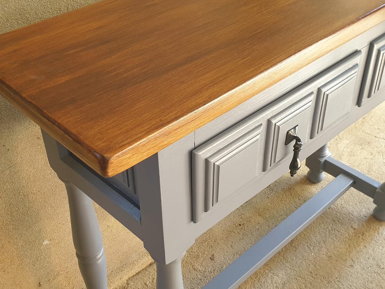 May include: A wooden console table with a stained brown top and a gray painted base. The table features three drawers with decorative molding and a keyhole. The legs are turned and the table has a sturdy, gray support beam.