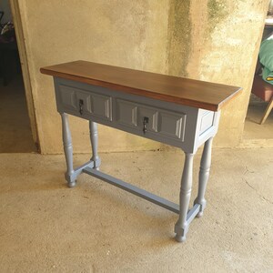 May include: A gray console table with a dark brown wooden top. The table has two drawers with black hardware and four gray, turned legs. The table is approximately 48 inches long.