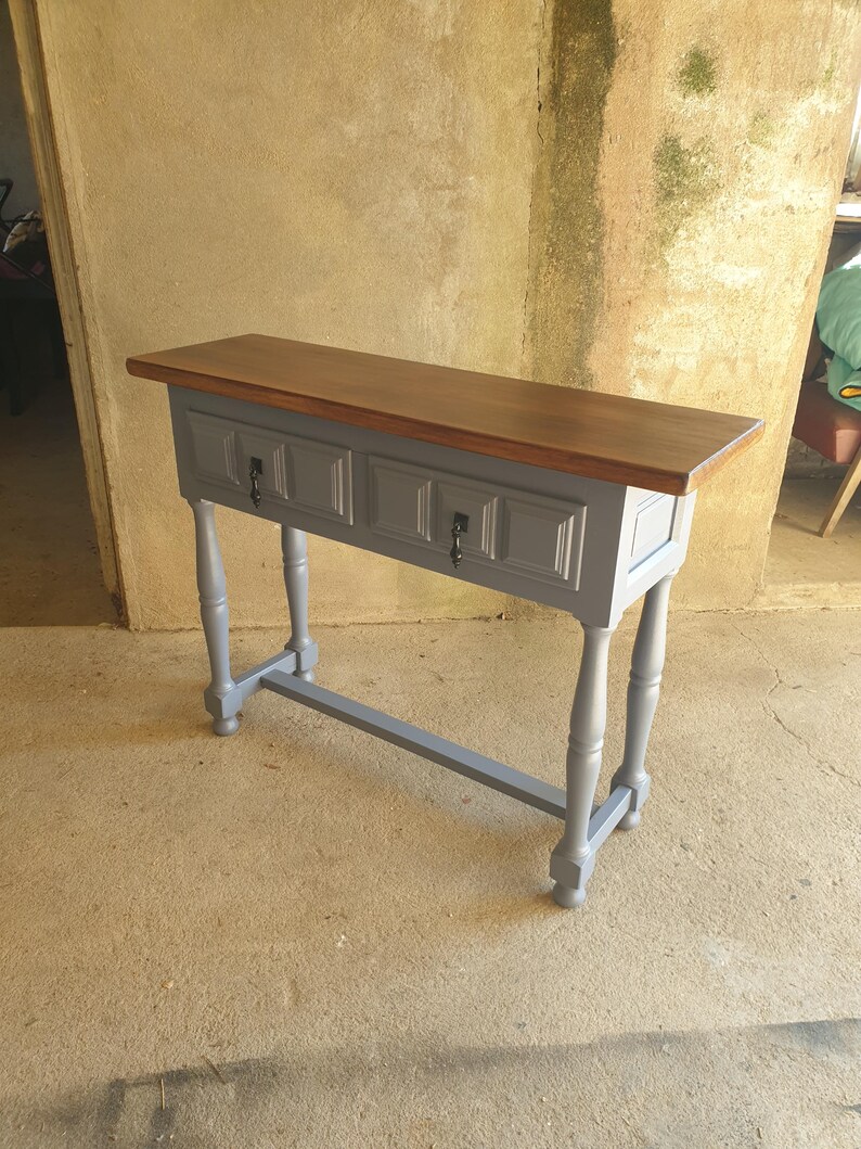 May include: A gray console table with a dark brown wooden top. The table has two drawers with black hardware and four gray, turned legs. The table is approximately 48 inches long.