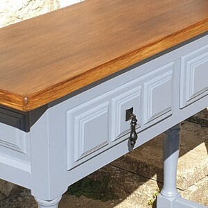 May include: A wooden console table with a light gray base and a dark brown top. The table features three drawers with decorative panels and ornate black metal handles. The legs are also light gray.