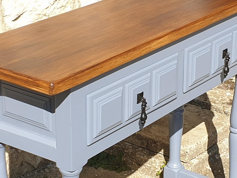 May include: A wooden console table with a light gray base and a dark brown top. The table features three drawers with decorative panels and ornate black metal handles. The legs are also light gray.