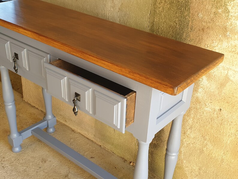 May include: A gray console table with a brown wooden top and two drawers. One drawer is open, revealing the interior. The table has ornate legs and drawer pulls.