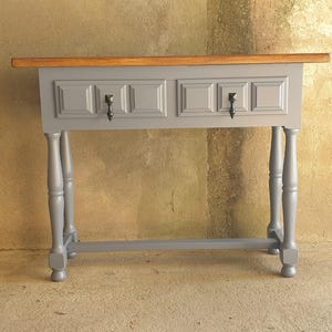 Peut inclure: Une console grise avec deux tiroirs et un dessus en bois. La table a des pieds tournés et une quincaillerie noire. Les tiroirs ont un design à panneaux encastrés. La table est peinte en gris mat.