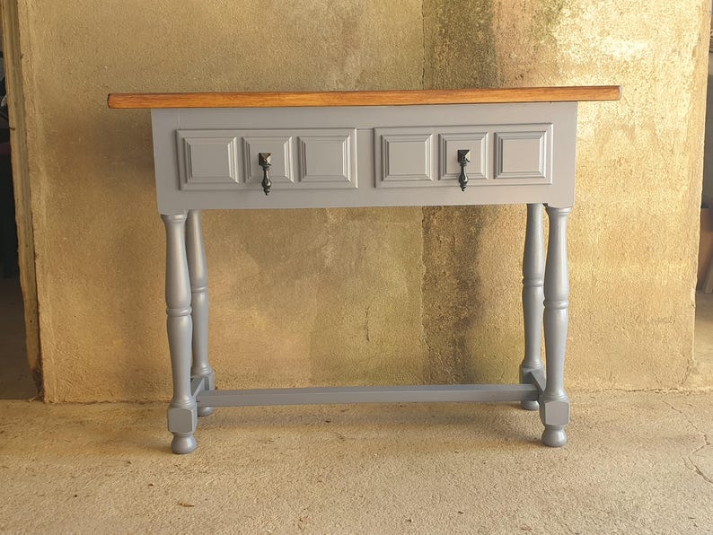 May include: A gray console table with two drawers and a wooden top. The table has turned legs and black hardware. The drawers have a recessed panel design. The table is painted in a matte gray finish.