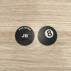 May include: Two circular black coasters. The left coaster features a textured surface with the white text PUB NAME and JB. The right coaster is smooth and displays a white circle containing the black number 8, resembling a billiard ball.