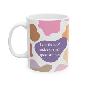 Puede incluir: Taza de cerámica blanca con asa, con un diseño colorido de estampado de vaca en tonos rosa, marrón y naranja. La taza tiene el texto "I can fix your underbite, not your attitude" en un bocadillo morado.