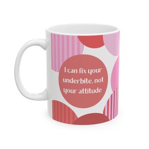 Puede incluir: Taza de cerámica blanca con asa curva. La taza presenta un diseño de círculos y rayas rosas y rojas. El texto de la taza dice: "I can fix your underbite, not your attitude."