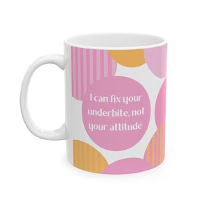 Puede incluir: Taza de cerámica blanca con asa, con un diseño de círculos y rayas rosas y naranjas. La taza tiene el texto: "I can fix your underbite, not your attitude."