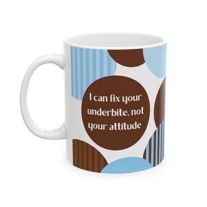 Puede incluir: Taza de cerámica blanca con asa, con un diseño de círculos y rayas azules y marrones. La taza tiene el texto "I can fix your underbite, not your attitude" en un círculo marrón. Un regalo divertido.
