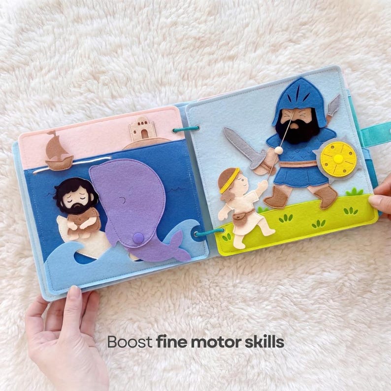 Pu&ograve; includere: Un libro di stoffa in feltro con illustrazioni di scene bibliche. Una pagina mostra un uomo in barca e una balena viola. L'altra pagina raffigura un guerriero con una spada e uno scudo. Il testo "Boost fine motor skills" &egrave; in basso.