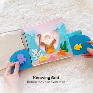 Pu&ograve; includere: Un libro tranquillo in feltro a tema biblico. Il libro aperto mostra una scena con un personaggio dei cartoni animati, onde e creature marine. Il testo "Knowing God before they can even read" &egrave; in basso.
