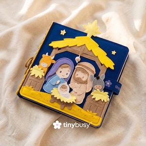 May include: A blue felt book with a yellow star and a nativity scene. The scene includes Mary, Joseph, baby Jesus, a donkey, and a cow. The book has a yellow border and a strap with the word "tiny" on it.