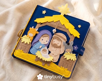 May include: A blue felt book with a yellow star and a nativity scene. The scene includes Mary, Joseph, baby Jesus, a donkey, and a cow. The book has a yellow border and a strap with the word "tiny" on it.