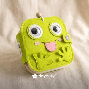 May include: A bright green felt frog-shaped book with large googly eyes, a pink tongue, and small felt hands. The book features a wooden bead on top and a purple flower clasp. The "tinybusy" logo is at the bottom.