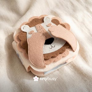 May include: A soft, fabric activity book shaped like a lion. The lion has a light brown mane, a white face, and tan paws. The book is closed, with the lion's face visible. The book is on a cream-colored fabric.