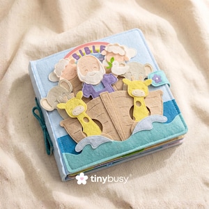 May include: A blue felt Bible storybook with a felt applique scene of Noah's Ark. The cover features a rainbow, clouds, and the word "BIBLE". The ark includes giraffes and other animals. The book is secured with a teal tie closure.