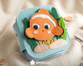 May include: A blue felt book featuring an orange clownfish design. The fish has white stripes and googly eyes, with green seaweed and coral accents. The book is closed with a button fastening. The book is approximately 15 cm square.