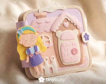 May include: A felt activity book featuring a house and a doll. The house is pink, tan, and lavender with a heart-shaped roof and a door with a paw print. The doll has blonde hair and wears a blue headband and purple dress. The book has the text "tinybusy".