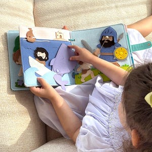 Pu&ograve; includere: Un libro di feltro con pi&ugrave; pagine. Una pagina mostra una scena con un uomo in barca, una balena e onde. Un'altra pagina raffigura un cavaliere con una spada e uno scudo. Il libro &egrave; tenuto aperto da un bambino.