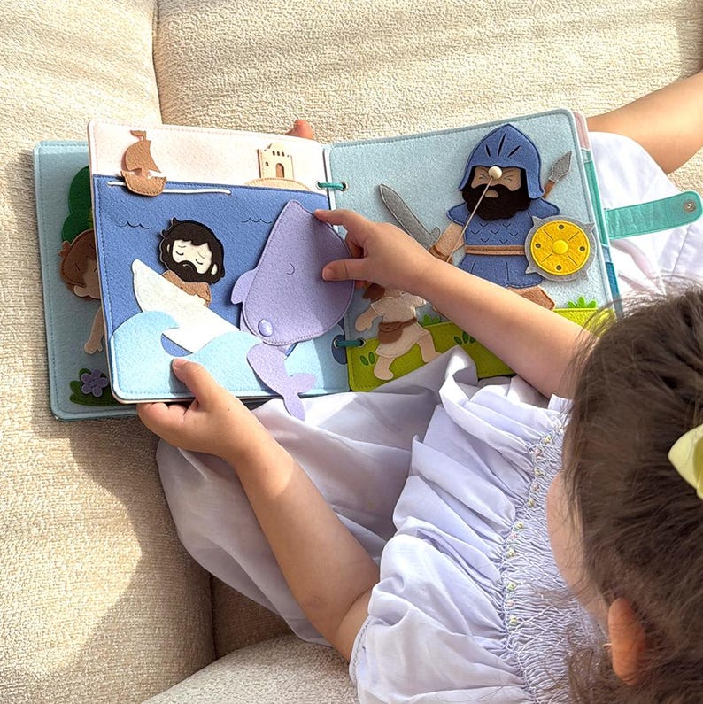 Pu&ograve; includere: Un libro di feltro con pi&ugrave; pagine. Una pagina mostra una scena con un uomo in barca, una balena e onde. Un'altra pagina raffigura un cavaliere con una spada e uno scudo. Il libro &egrave; tenuto aperto da un bambino.