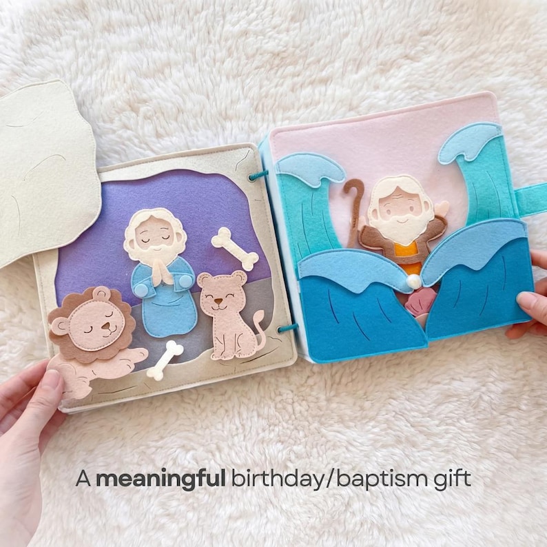 Pu&ograve; includere: Un libro di stoffa in feltro con scene di una storia biblica. Il libro aperto mostra un leone, un orso e un uomo in una grotta da un lato, e un uomo nell'acqua dall'altro. Il testo in basso dice "A meaningful birthday/baptism gift."