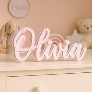 Può includere: Un'insegna con il nome "Olivia" in corsivo, rosa e bianco a strati, esposta su una superficie bianca. L'insegna è il punto focale, con uno sfondo rosa tenue e un coniglietto, un orsacchiotto e una stella sullo sfondo.