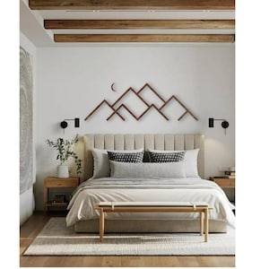 Może przedstawiać: Sypialnia z neutralną paletą kolorów. Drewniana dekoracja ścienna w kształcie gór wisi nad łóżkiem z beżowym wezgłowiem. Łóżko jest pościelone białą i szarą pościelą, a u jego podnóża stoi drewniana ławka.