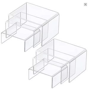 Könnte beinhalten: Set aus drei transparenten Acryl-Display-Erhöhungen, die zur platzsparenden Aufbewahrung ineinander gesteckt werden können. Jede Erhöhung hat eine rechteckige Oberseite und eine U-förmige Basis. Das transparente Design ermöglicht eine optimale Sichtbarkeit der ausgestellten Artikel.