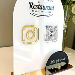 Może przedstawiać: Biały szyld restauracji ze złotymi akcentami. Szyld zawiera napis "Restaurant Tradicional Food" oraz logo Instagram. Obecny jest również kod QR. Podstawa szyldu jest czarna z napisem "Let's get social".