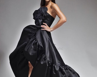 May include: A strapless black gown with a high-low hemline and a large floral accent on the shoulder. The dress features a flowing skirt and is paired with gold heels. The overall style is elegant and formal.