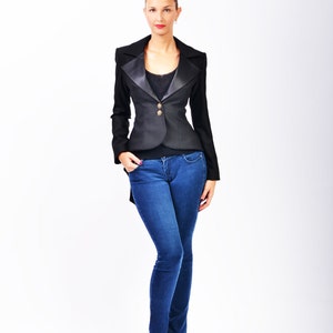 May include: A woman wearing a black blazer with a satin lapel and blue jeans. She is standing with her hands in her pockets.