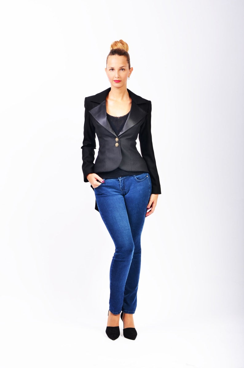 May include: A woman wearing a black blazer with a satin lapel and blue jeans. She is standing with her hands in her pockets.