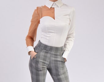 May include: A white long-sleeved blouse with a sheer brown overlay on one sleeve. The blouse has a unique design with a heart-shaped cutout at the front. The model is wearing grey checked trousers.