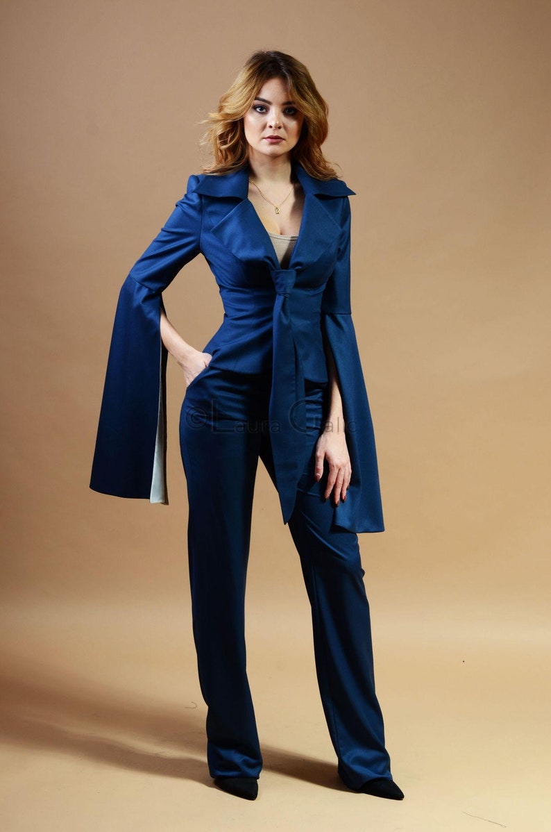 May include: A woman wearing a dark blue pantsuit with a long, flowing jacket and wide sleeves. The jacket has a tie at the waist and the pants are straight-legged.