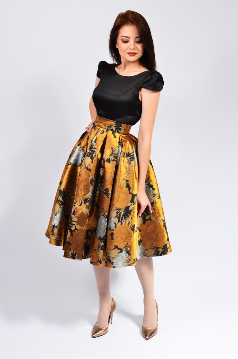 May include: A woman wearing a black top and a gold and black floral skirt. She is wearing gold heels.