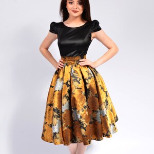 May include: A woman wearing a black satin top with a gold and black floral skirt. She is standing on a white background and wearing gold heels.
