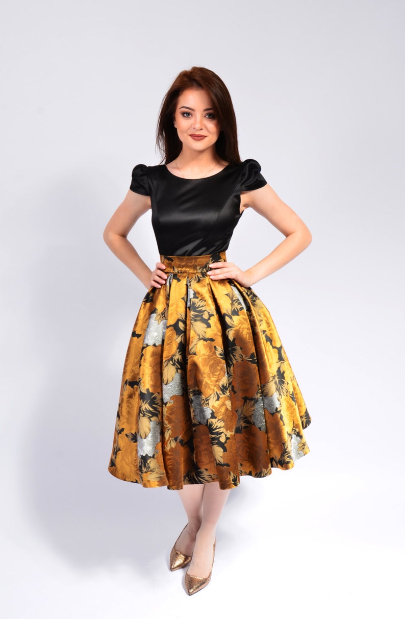 May include: A woman wearing a black satin top with a gold and black floral skirt. She is standing on a white background and wearing gold heels.